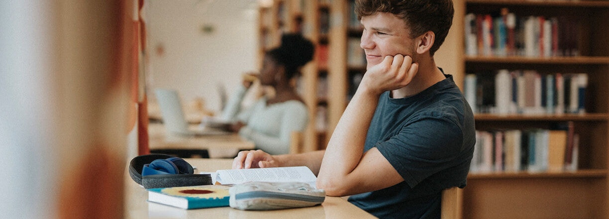 Um estudante a estudar numa secretária com livros e um portátil numa biblioteca.