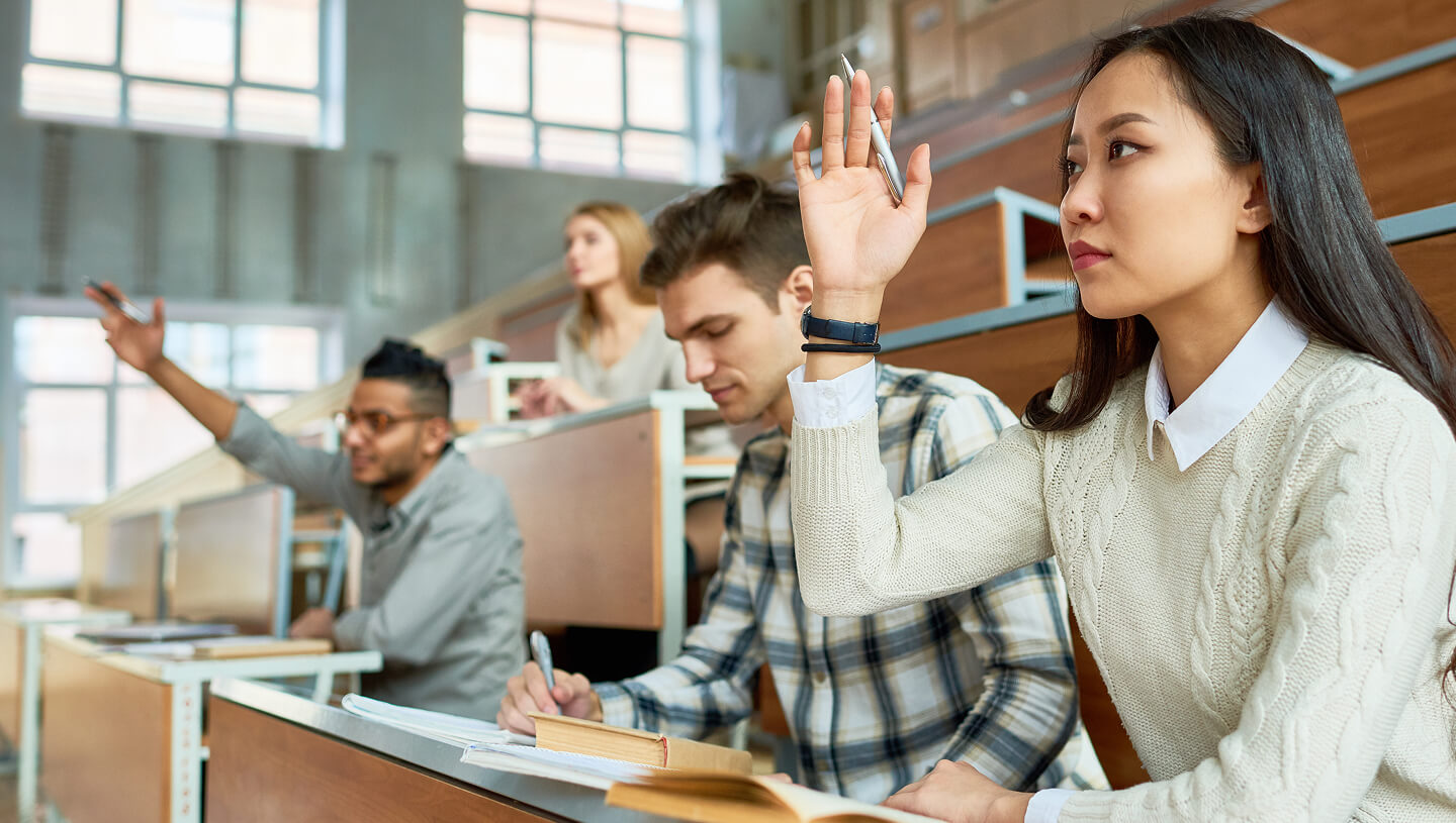 Uma sala de aula universitária com estudantes sentados em secretárias, enquanto uma estudante em primeiro plano levanta a mão.